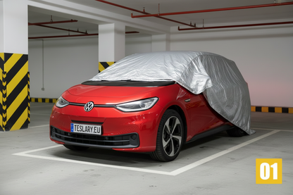 A red Volkswagen electric car parked in an indoor parking garage, with a silver car cover draped over the roof and side. The car features a license plate reading TESLARY-EU, and a yellow '01' badge is in the bottom right corner.
