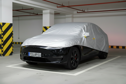 A black Tesla Model Y is parked inside a modern parking structure, covered with a metallic silver protective car cover. The vehicle's front license plate reads TESLARY EU.